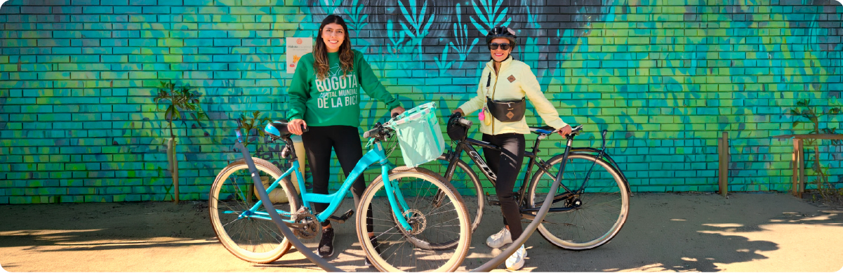 Fotografía de dos mujeres junto a sus bicicletas frente a un mural urbano de tonos verdes y azules. La imagen representa la movilidad sostenible en Bogotá, el uso de la bicicleta como medio de transporte y la participación ciudadana en iniciativas que promueven formas de movilidad activa y sostenibles en la ciudad.