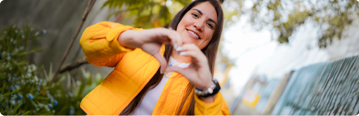 Fotografía de una mujer sonriente con chaqueta amarilla que forma un corazón con sus manos en un espacio al aire libre. La imagen representa a las mujeres como grupo poblacional y su participación activa en espacios ciudadanos, sociales y comunitarios de Bogotá.
