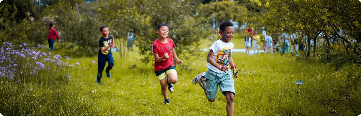 Fotografía de varios niños corriendo y jugando al aire libre en una zona verde rodeada de árboles y vegetación. La imagen representa a los niños, niñas y adolescentes como grupo poblacional y su derecho a participar, expresarse y disfrutar de espacios de encuentro, convivencia y desarrollo en Bogotá.