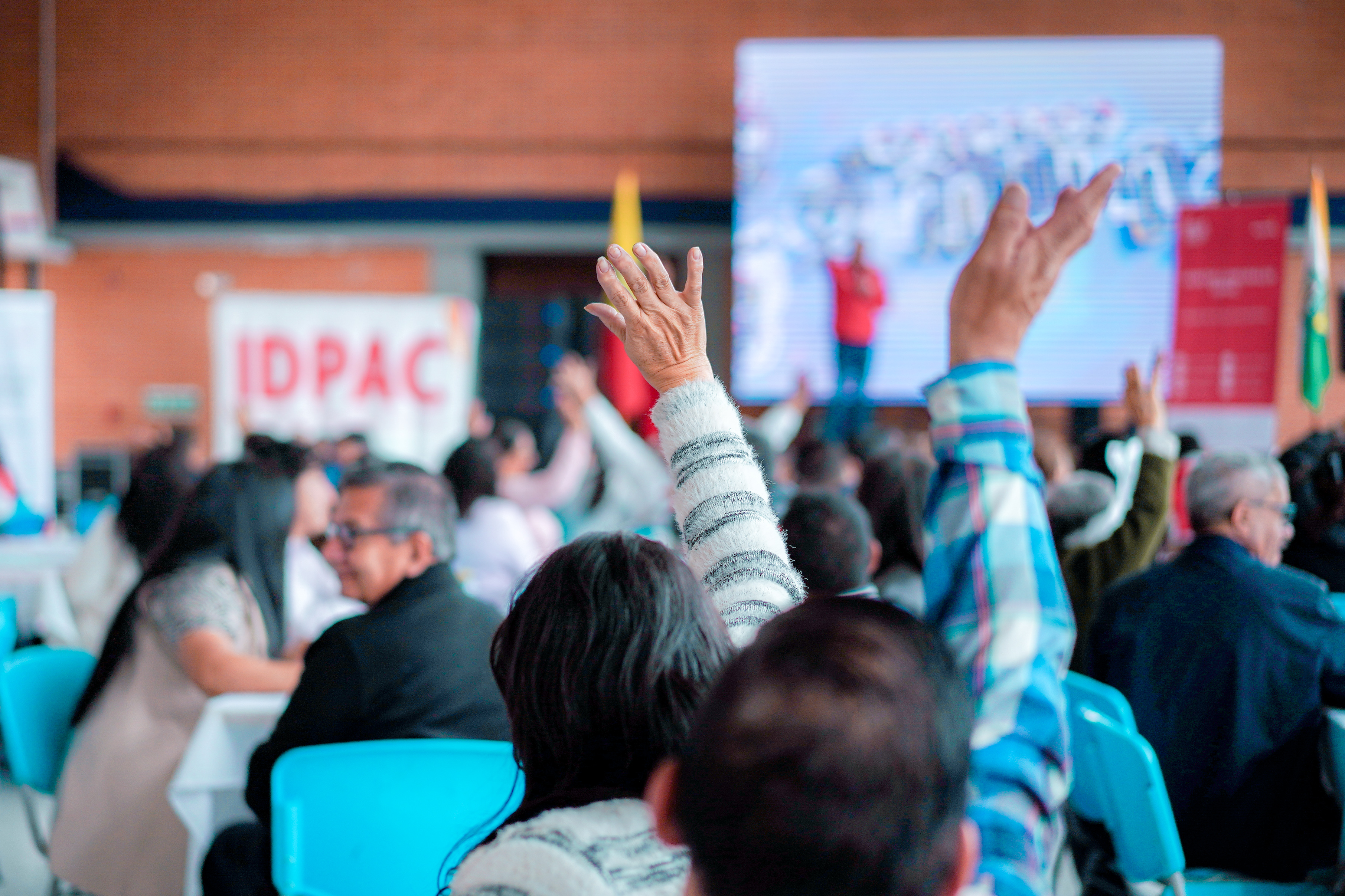 Ciudadanos levantan la mano para participar durante un evento del IDPAC en Bogotá, con pantalla y pendón institucional de fondo.