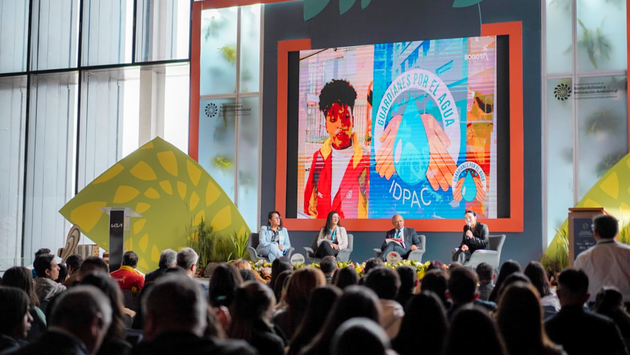 Panelistas del IDPAC participan en el conversatorio “Guardianes por el Agua” durante la Cumbre Internacional de Sostenibilidad, con una pantalla al fondo que promueve el cuidado del agua y la acción ciudadana en Bogotá