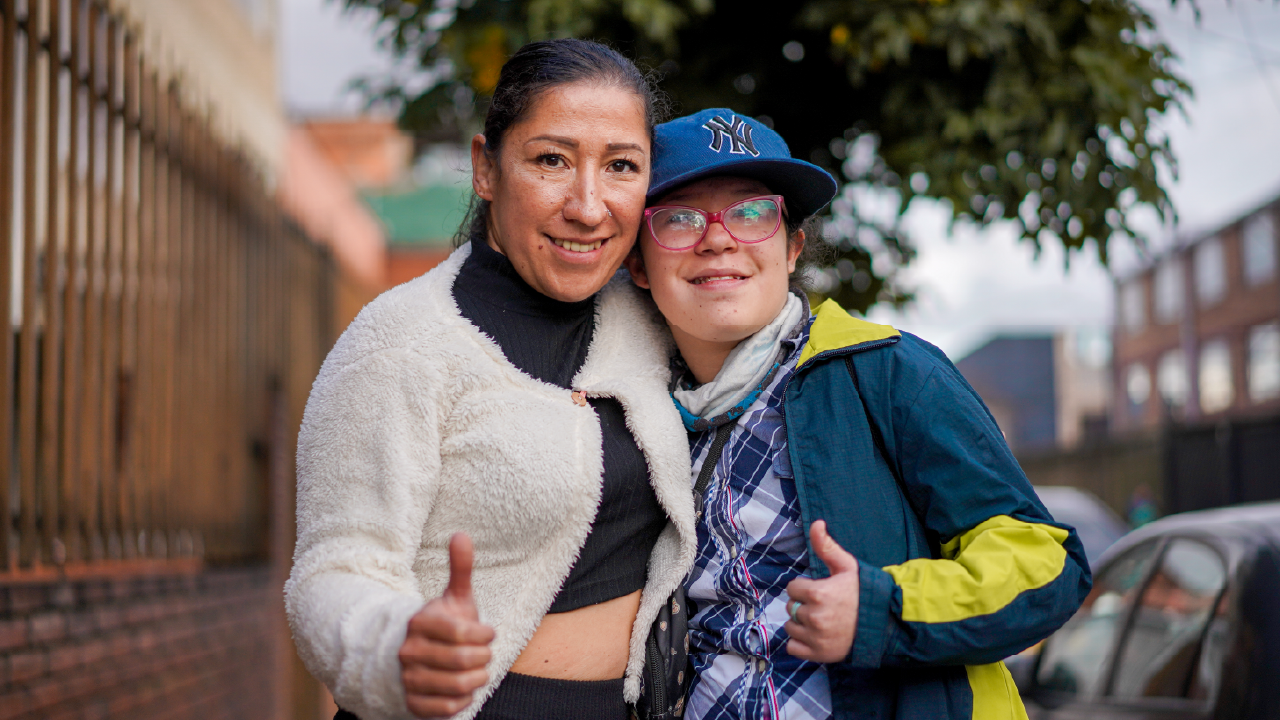 Dos personas sonríen y hacen pulgar arriba, abrazadas en una calle; retrato de inclusión y apoyo.