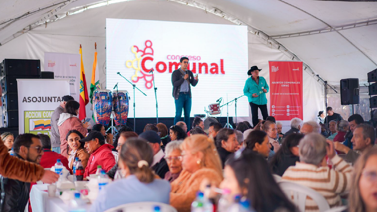 Líderes y comunidad participan en un Congreso Comunal Local en Bogotá, con escenario principal, panelistas y mesas de trabajo bajo una carpa.