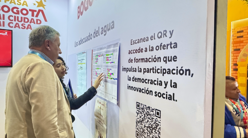 Persona explica a un visitante el ejercicio sobre el uso adecuado del agua en el stand del IDPAC durante la Cumbre de Sostenibilidad, destacando la oferta formativa en participación, democracia e innovación social.