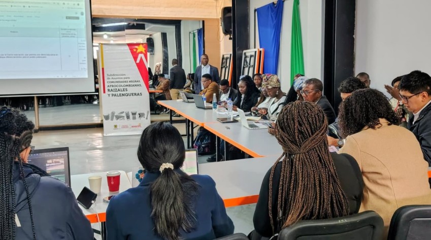Mesa de trabajo con delegados y delegadas de comunidades negras, afrocolombianas, raizales y palenqueras reunidos en jornada de concertación para revisar avances del CONPES 39, en un salón con proyector y banner institucional del IDPAC