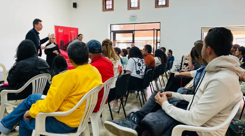 Personas participando en un encuentro comunitario del IDPAC dentro de un salón, escuchando a un facilitador al frente.