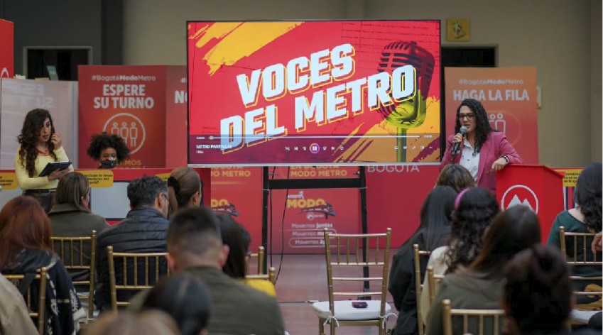 Presentadora habla al público durante el lanzamiento del pódcast 'Voces del Metro', proyectado en pantalla, en un evento de la estrategia Bogotá Modo Metro.
